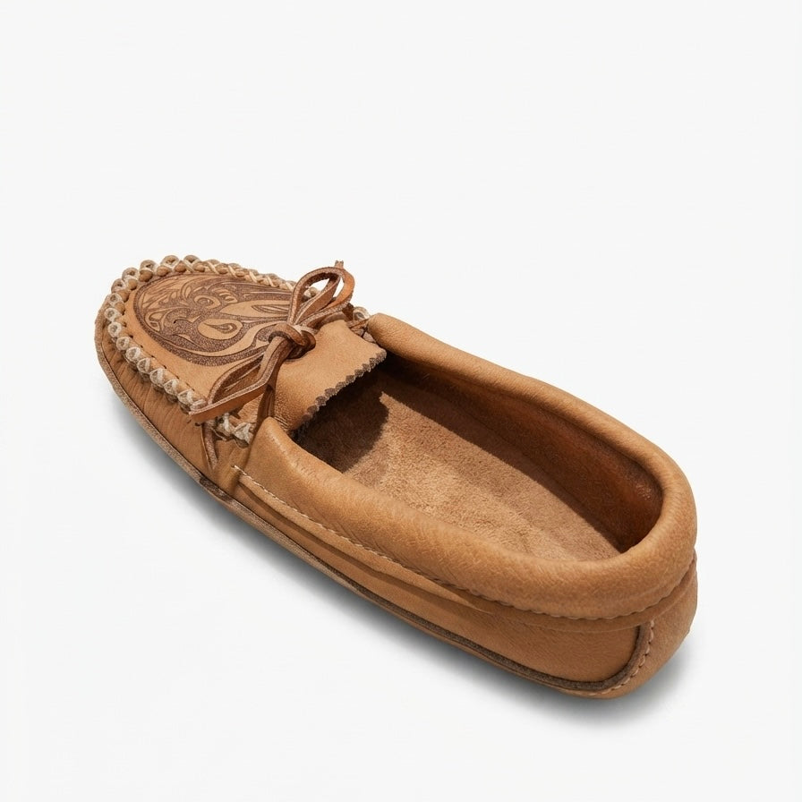 A single tan suede moccasin slipper with a braided trim and a decorative embroidered patch on the vamp, shown from a three-quarters angle on a light gray background.