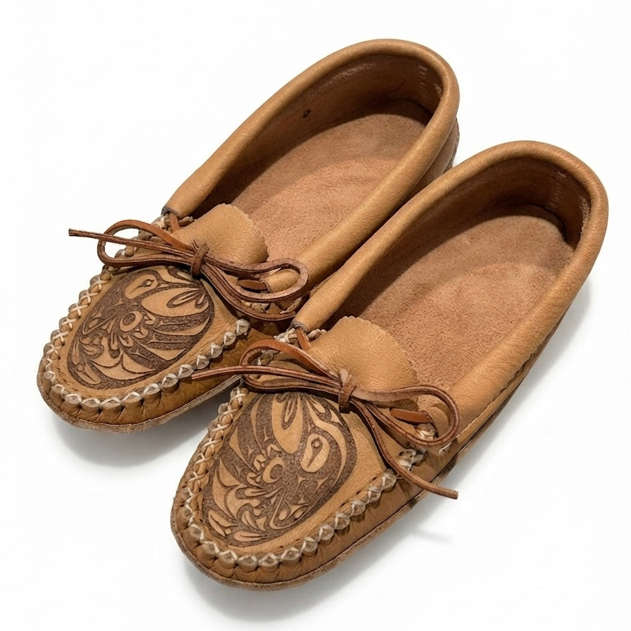A pair of tan suede moccasin-style slip-on shoes with hand-tooled floral detailing on the toes, braided trim, and matching leather bows, shown on a white background.