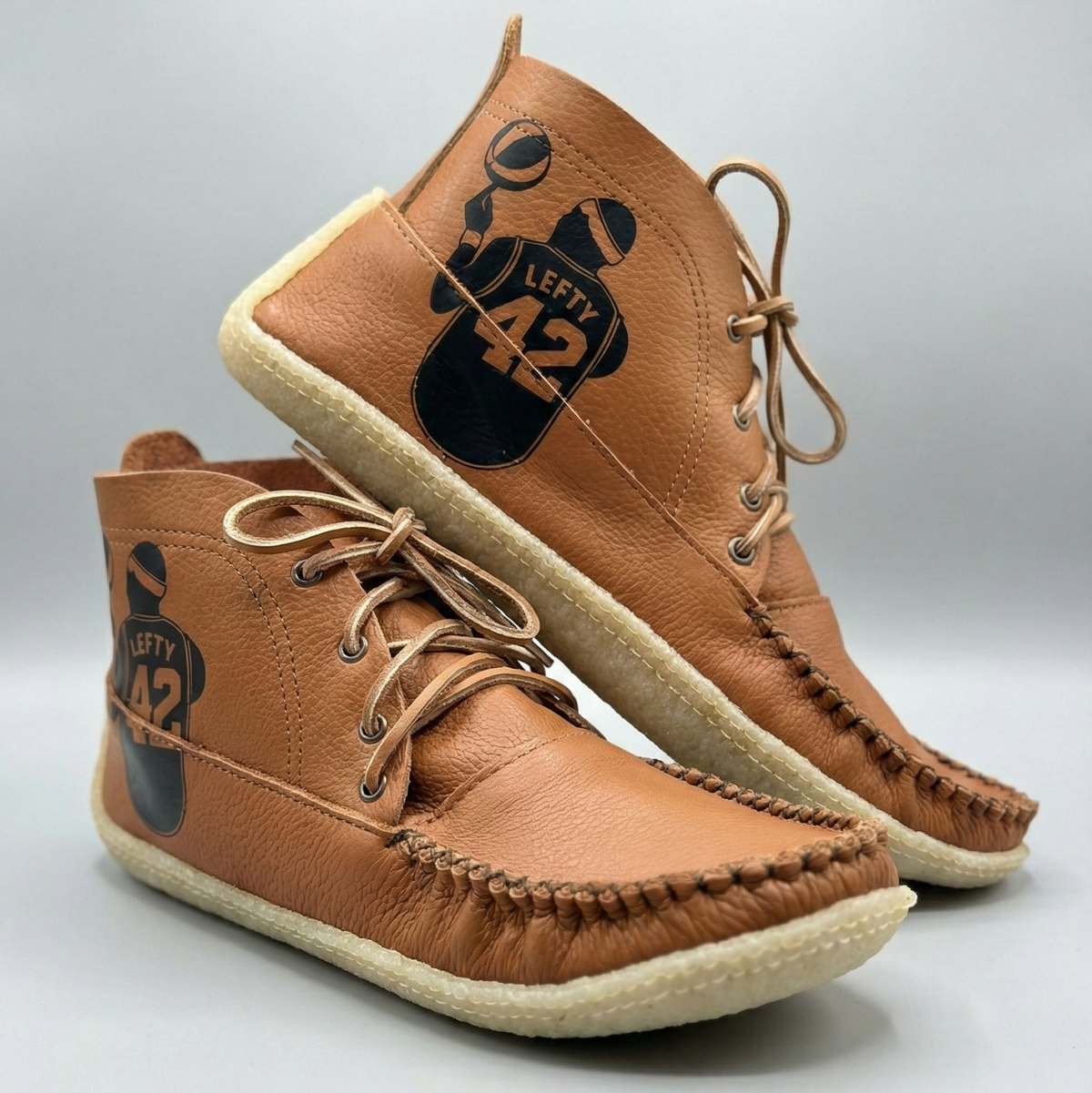A pair of dark tan leather moccasin-style mid-height lace-up shoes with cream soles and a black printed graphic on the outer side featuring a football player and the number 42.